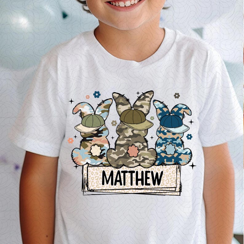 Puede incluir: Camiseta blanca con un gr&aacute;fico de tres conejos de Pascua de camuflaje con gorras de b&eacute;isbol. Los conejos est&aacute;n de pie sobre una pancarta de estampado de leopardo con el nombre "MATTHEW" escrito en negro.
