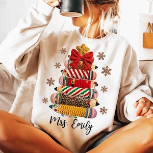May include: A white sweatshirt featuring a Christmas tree design made of stacked pencils. The pencils have various patterns, including red and white stripes, green and white stripes, and leopard print. A red bow and gold star top the tree, with snowflakes and the name "Mrs. Emily" below.