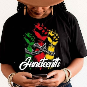 May include: Black t-shirt with a graphic of three fists in green, red, and yellow, linked by a chain. The word "Juneteenth" is written in white script below. The shirt is worn by a person.