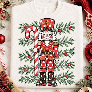 May include: A white crewneck sweatshirt featuring a graphic of a nutcracker wearing a red and white polka dot uniform. The nutcracker holds a large red and white striped candy cane. The background design includes green pine branches and red berries.