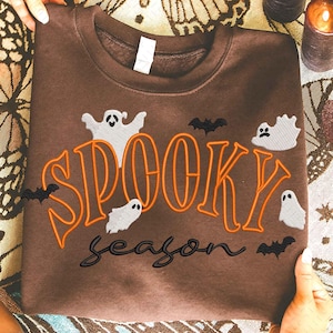 May include: A brown sweatshirt featuring the phrase "Spooky Season" in orange and black. The design includes white ghost and black bat graphics, ideal for Halloween.