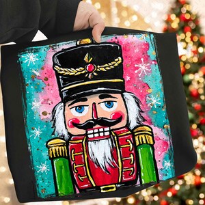 May include: Black tote bag featuring a colorful illustration of a nutcracker soldier. The nutcracker has a black hat with gold trim, a red uniform, and a white beard. The background is a mix of pink, teal, and gold accents, with snowflakes.