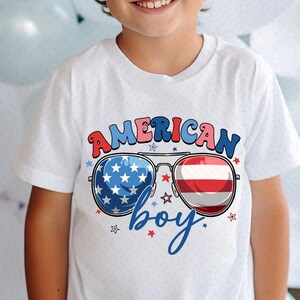 May include: White t-shirt with the words "AMERICAN boy" in red, white, and blue. The design includes sunglasses with the American flag and red and white stripes. Stars surround the text and sunglasses.