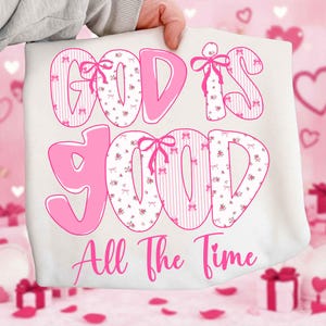 May include: A white tote bag with the phrase "GOD IS GOOD All The Time" in pink. The letters are outlined in pink and filled with floral and striped patterns, accented with pink bows.