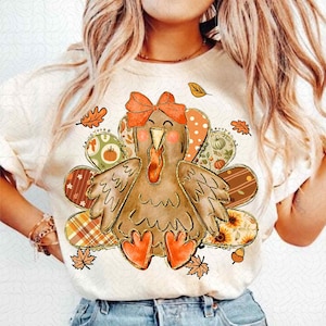 Könnte beinhalten: Cremefarbenes T-Shirt mit einer Cartoon-Truthahn-Illustration, verziert mit einer Schleife und Herbstblättern. Die Schwanzfedern des Truthahns weisen verschiedene Muster auf, darunter Kürbisse, Sonnenblumen und Tartan. Perfekt für Thanksgiving.