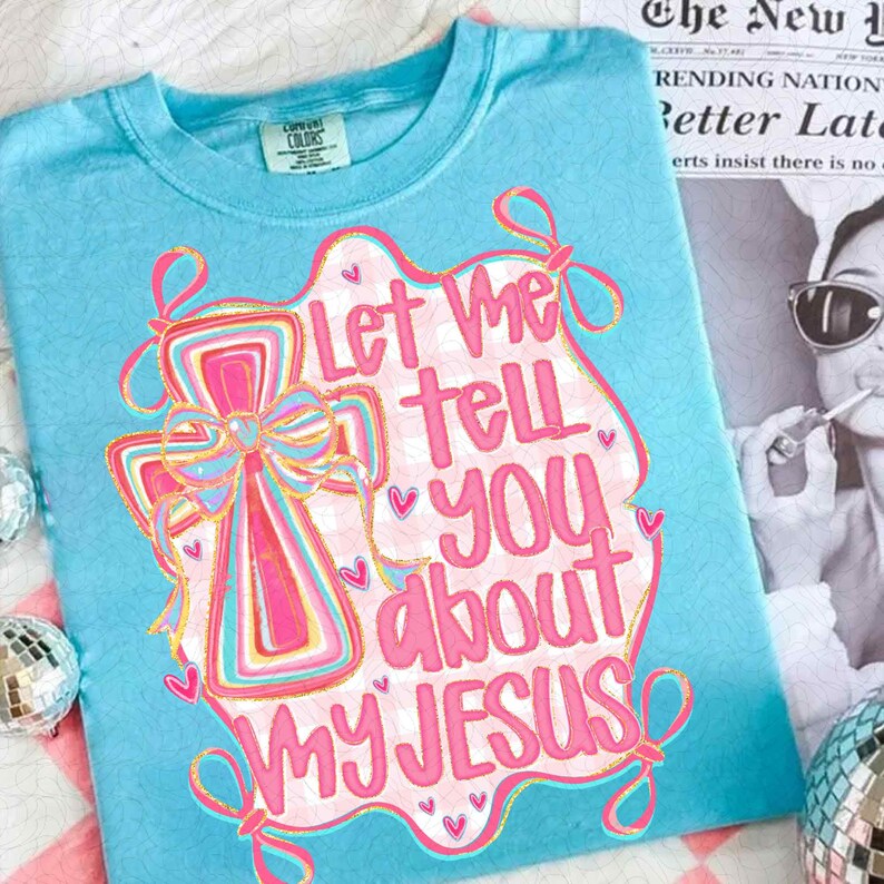 K&ouml;nnte beinhalten: Hellblaues T-Shirt mit dem Text "Let Me Tell You About My Jesus" in rosa Schrift. Das Design beinhaltet ein Kreuz mit Schleife und Herzen. Das Shirt ist aus weichem Material.