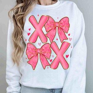 May include: White sweatshirt with a pink X and bow design. The bows have a heart pattern and gold trim. Red hearts and stars are scattered around the design. A casual top for Valentine's Day or everyday wear.