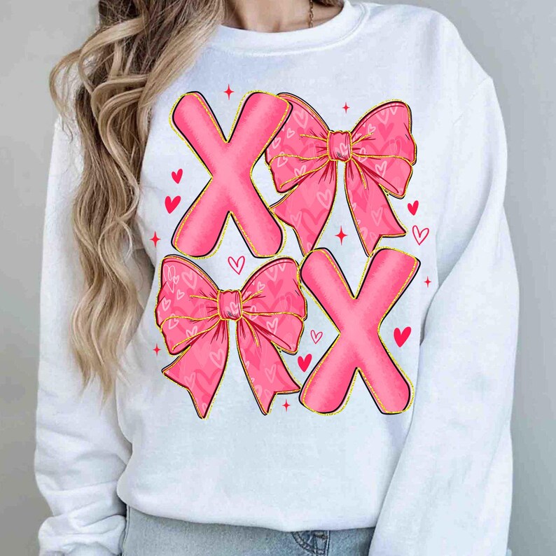 May include: White sweatshirt with a pink X and bow design. The bows have a heart pattern and gold trim. Red hearts and stars are scattered around the design. A casual top for Valentine's Day or everyday wear.
