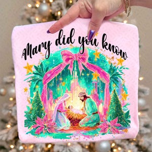 May include: A pink, square-shaped item with the text "Mary did you know" in black script. The image features a Nativity scene with Mary, Joseph, and baby Jesus, surrounded by a decorative arch and stars. The design includes pink, green, and gold accents.