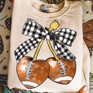 May include: A beige sweatshirt featuring a graphic of two footballs shaped like cherries, with white stitching and a black and white checkered bow. The stems are gold. The design is centered on the front of the shirt.