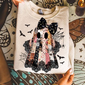 May include: Beige sweatshirt featuring a patchwork ghost design with a black bow, lace trim, and bat and spiderweb accents. The ghost is made of various floral and patterned fabrics, perfect for Halloween.