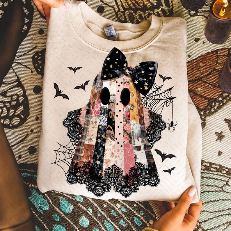 May include: Beige sweatshirt featuring a patchwork ghost design with a black bow, lace trim, and bat and spiderweb accents. The ghost is made of various floral and patterned fabrics, perfect for Halloween.