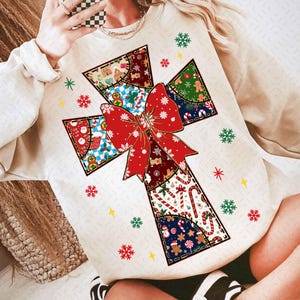 Puede incluir: Sudadera de color crema con un gran diseño de cruz de patchwork con un lazo rojo. La cruz está compuesta por varios patrones con temática navideña, incluyendo hombres de jengibre, bastones de caramelo y copos de nieve. Acentos festivos incluyen copos de nieve y estrellas.