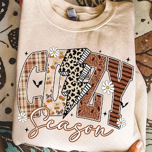 May include: Beige sweatshirt with a colorful graphic that says "Cozy Season." The letters are decorated with plaid, leopard print, and fall-themed images. The design includes daisies and bats.
