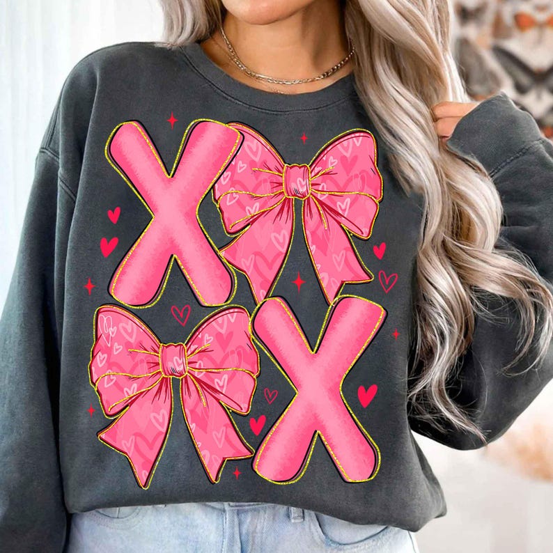 May include: Dark gray sweatshirt featuring a pink "XOXO" design. The letters are outlined in gold and adorned with pink bows decorated with heart patterns. Small red hearts and stars are scattered around the letters, creating a Valentine's Day theme.