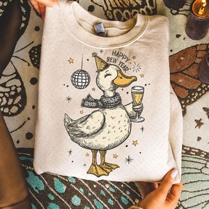 May include: Cream-colored sweatshirt featuring a cartoon duck wearing a party hat and scarf, holding a champagne glass. The duck is surrounded by stars and a disco ball, with "HAPPY NEW YEAR" text. A festive design for the New Year.