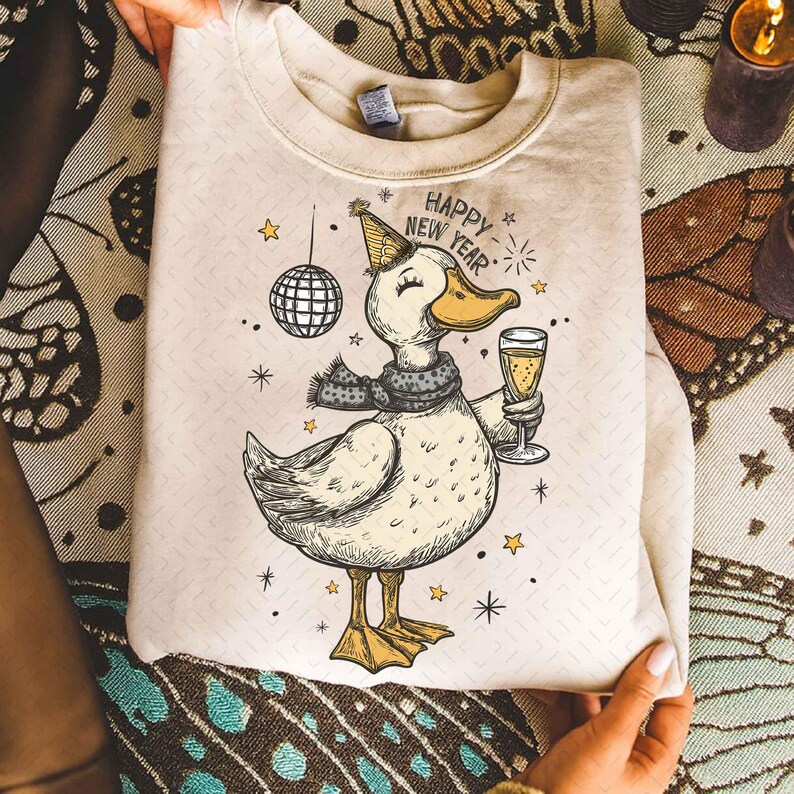 May include: Cream-colored sweatshirt featuring a cartoon duck wearing a party hat and scarf, holding a champagne glass. The duck is surrounded by stars and a disco ball, with "HAPPY NEW YEAR" text. A festive design for the New Year.
