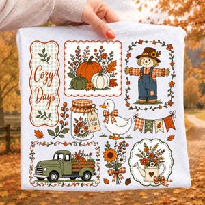 May include: A white t-shirt featuring a colorful autumn-themed graphic print. The design includes a scarecrow, a green truck, a duck, pumpkins, a mason jar with a tag reading Hello Fall, a watering can with flowers, and the text Cozy Days.