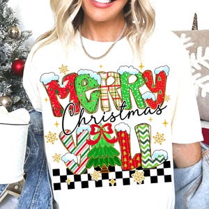 May include: Cream-colored t-shirt with a festive "Merry Christmas Y'all" graphic. The text features red, green, and white patterns with snow accents. A Christmas tree and snowflakes complete the holiday design.