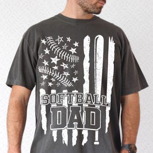 Op de afbeelding: Donkergrijs t-shirt met een wit grafisch ontwerp dat op een vlag lijkt. Het ontwerp bevat een honkbal, sterren en de woorden "SOFTBALL DAD" in vette letters. Het shirt heeft korte mouwen en een relaxte pasvorm.