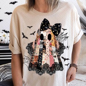 May include: Beige t-shirt featuring a patchwork ghost design with a black bow adorned with gold stars. The ghost has large black eyes and is decorated with lace and floral patterns. Black bats and a spiderweb complete the Halloween theme.