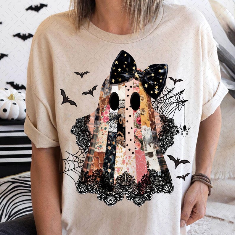 May include: Beige t-shirt featuring a patchwork ghost design with a black bow adorned with gold stars. The ghost has large black eyes and is decorated with lace and floral patterns. Black bats and a spiderweb complete the Halloween theme.