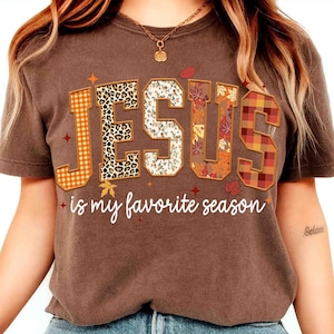 May include: Brown t-shirt with the text "JESUS is my favorite season." The word "JESUS" is designed with various patterns, including leopard print, floral, and plaid. The shirt has a crew neck and short sleeves.