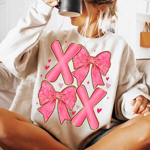 May include: Cream-colored sweatshirt featuring a pink "XOXO" design. The design includes two pink "X" letters and two pink bows with heart patterns. Small red hearts and stars are scattered around the design.