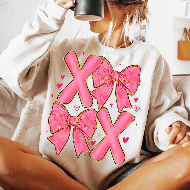 May include: Cream-colored sweatshirt featuring a pink "XOXO" design. The design includes two pink "X" letters and two pink bows with heart patterns. Small red hearts and stars are scattered around the design.