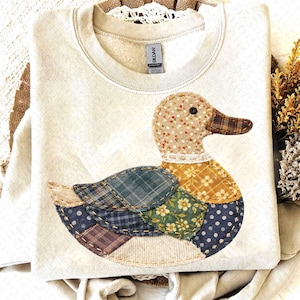 May include: Cream-coloured sweatshirt with a patchwork duck design. The duck has a patterned head, a brown beak, and wings made of various fabric patterns in blue, green, and brown. The sweatshirt has a crew neck and a tag that says "GILDAN".