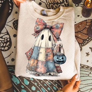 May include: Cream-colored sweatshirt featuring a whimsical ghost illustration. The ghost is adorned with a patchwork dress, a large floral bow, and holds a blue jack-o'-lantern bucket. The design has a vintage, hand-drawn aesthetic, perfect for Halloween.