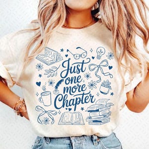 以下が含まれることがあります： 本、メガネ、そして「Just one more Chapter」の文字が描かれた、青いグラフィックデザインのクリーム色のTシャツ。デザインにはコーヒーカップ、リボン、花の要素が含まれており、文学的なテーマを作り出しています。