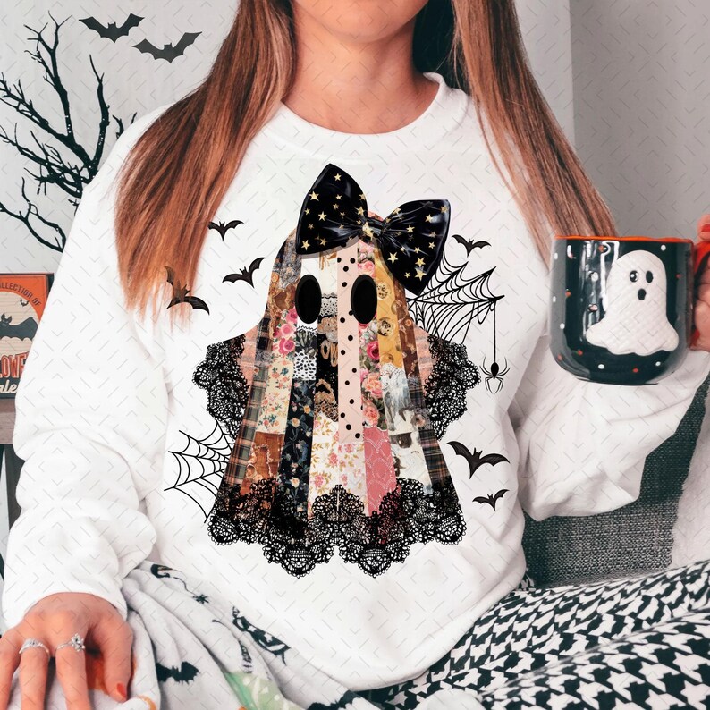 May include: White sweatshirt featuring a patchwork ghost design with a black bow, lace trim, and bat and spiderweb accents. A black and white ghost mug is held in the hand. Halloween-themed apparel.