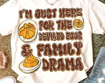 May include: White t-shirt with the text "I'M JUST HERE FOR THE DEVILED EGGS & FAMILY DRAMA" in brown and orange lettering. The shirt features illustrations of deviled eggs.