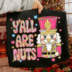 May include: Black tote bag with the text "Y'ALL ARE NUTS" in colorful bubble letters, surrounded by stars. A pink and gold nutcracker soldier is featured on the right side of the bag, with a gold crown and a white beard.