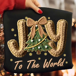 Può includere: Cuscino nero con la parola "JOY" in stile uncinetto, un albero di Natale all'uncinetto e la frase "To The World". L'albero ha ornamenti bianchi e un fiocco marrone. Fiocchi di neve dorati sono sparsi intorno al testo.