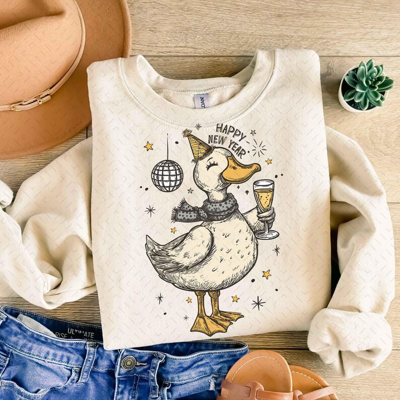 May include: Cream-colored sweatshirt with a graphic of a duck wearing a party hat, scarf, and holding a champagne glass. The text "Happy New Year" is above the duck. A disco ball and stars are also in the design.