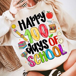 May include: Cream-colored sweatshirt with a colorful graphic celebrating 100 days of school. The design includes the words "Happy 100 Days of School" with images of an apple, pencil, books, and crayons.