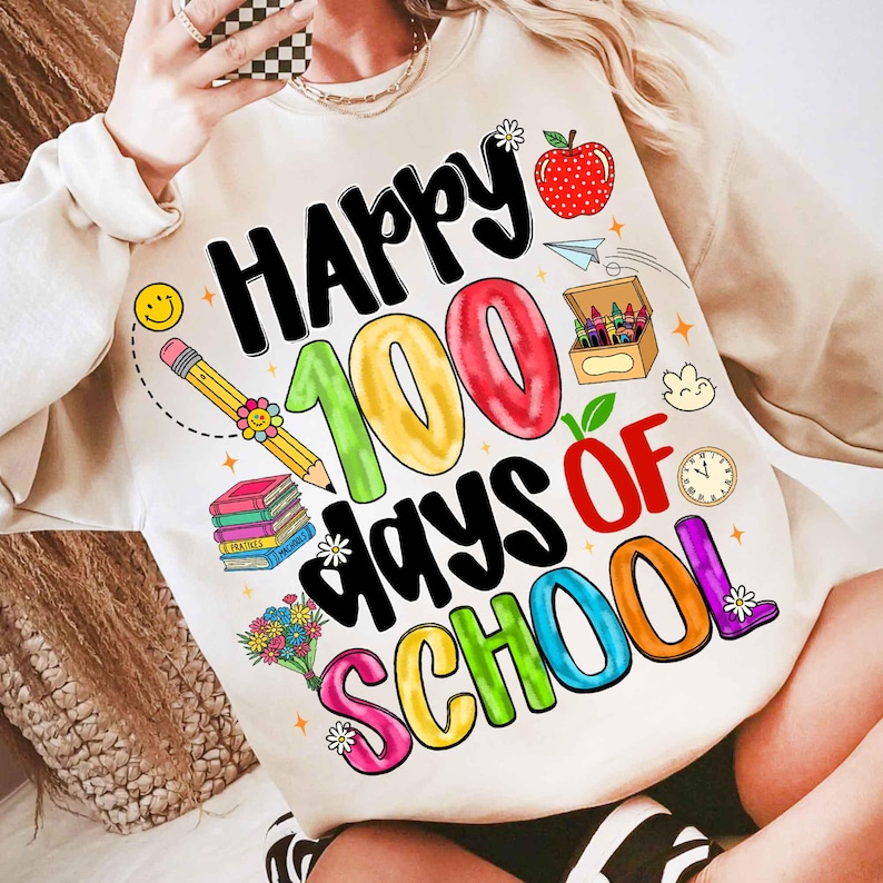May include: Cream-colored sweatshirt with a colorful graphic celebrating 100 days of school. The design includes the words "Happy 100 Days of School" with images of an apple, pencil, books, and crayons.