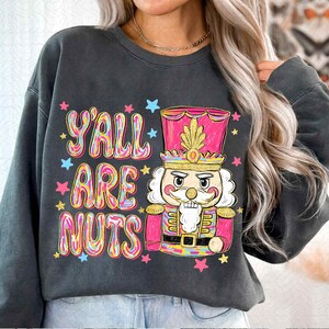 May include: Dark gray sweatshirt with a colorful graphic of a nutcracker and the text "Y'ALL ARE NUTS". The nutcracker is in pink, gold, and blue, with stars around the text. A festive holiday-themed design.