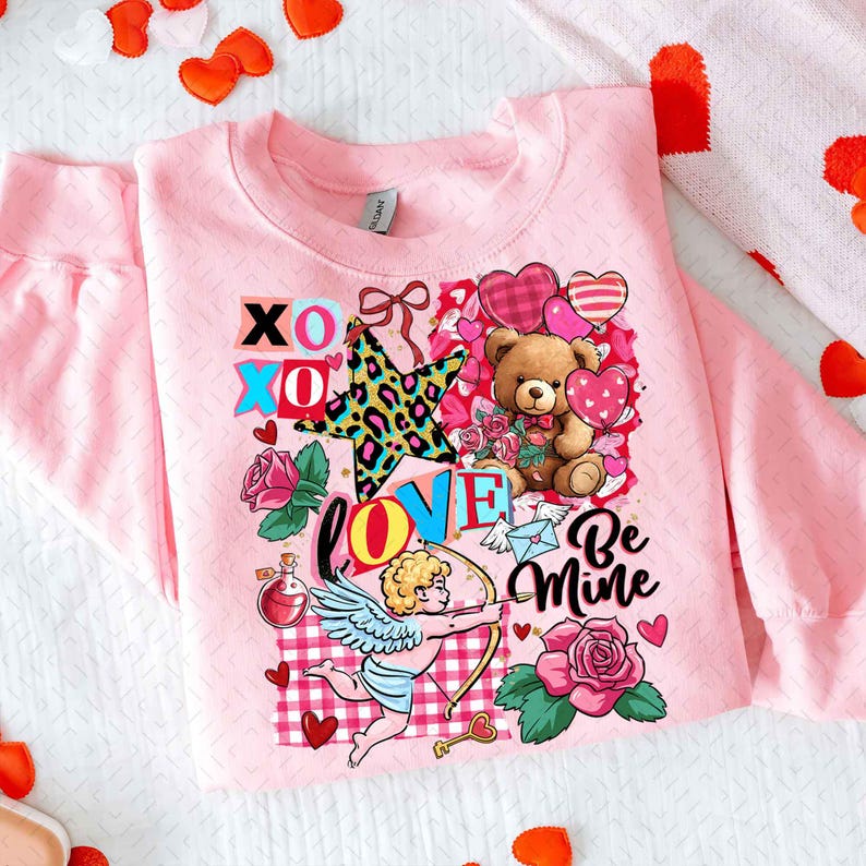 Op de afbeelding: Zachtroze sweatshirt met een Valentijnsdag-ontwerp. Het ontwerp bevat een teddybeer, harten, rozen en de woorden "XOXO", "LOVE" en "Be Mine". Een ster met een luipaardprint is ook te zien.