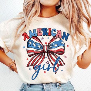 May include: Cream-colored t-shirt featuring a patriotic bow design with red, white, and blue colors. The text "AMERICAN" is arched above the bow, and "girl" is written in cursive below. Small stars are scattered around the bow.