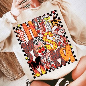 Peut inclure: Sweat-shirt crème avec un motif coloré « Hot Miss Mama ». Le texte utilise divers motifs, dont léopard, damier et cerises. Le design est encadré d'une bordure à carreaux noirs et blancs.