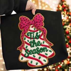 May include: Black tote bag featuring a Christmas bell design with a pink bow and the text "Tis the Season" in green. The bell is decorated with red and green stripes and dots, and a brown border.
