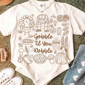May include: White t-shirt with a brown line drawing of Thanksgiving-themed images, including a turkey, scarf, leaves, and pie. The text "Gobble Til You Wobble" is also featured on the shirt. A silver necklace is visible at the neckline.