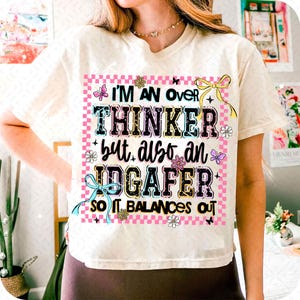 May include: Cream-colored t-shirt with the text "I'M AN OVER THINKER but also an IDGAFER so it balances out" in a colorful, playful font. The design includes butterflies, flowers, and bows.
