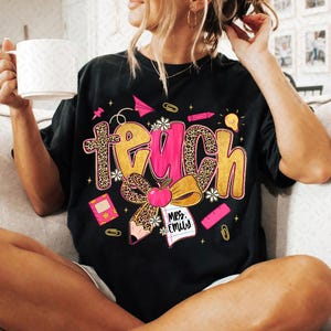 May include: Black t-shirt with the word "Teach" in a pink and leopard print design. The shirt features school-themed graphics like an apple, pencil, and notebook. The text "Mrs. Emily" is also visible.