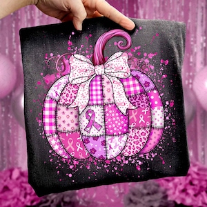 May include: A black pillow with a pink pumpkin design. The pumpkin is made of patchwork with various pink patterns, including ribbons, flowers, and leopard print. A large bow and stem top the pumpkin. Pink paint splatters the background.