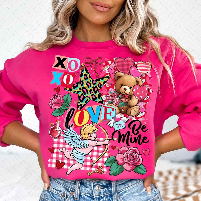 Op de afbeelding: Een felroze sweatshirt met een Valentijnsdagontwerp. Het ontwerp bevat een teddybeer, harten, rozen, een ster en de woorden "LOVE" en "Be Mine". Er is ook een cupido met pijl en boog afgebeeld.