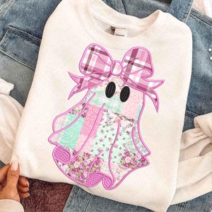 May include: White sweatshirt featuring a ghost design with a pink plaid bow and patchwork body. The ghost's body is composed of floral and patterned fabrics in shades of pink and mint green. Black oval eyes complete the design.